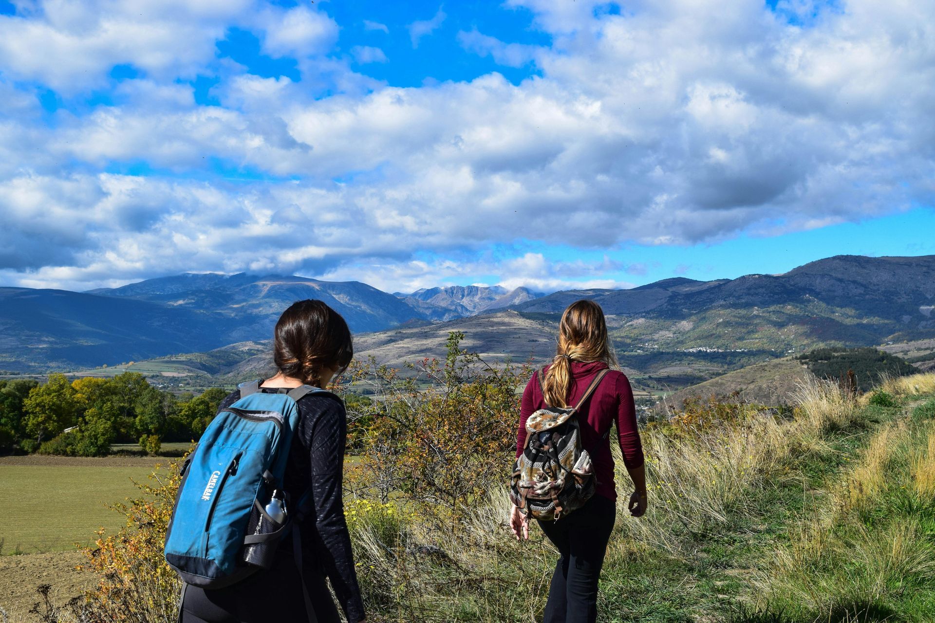 Two women hike on a path with mountains in the background, under a partly cloudy blue sky.