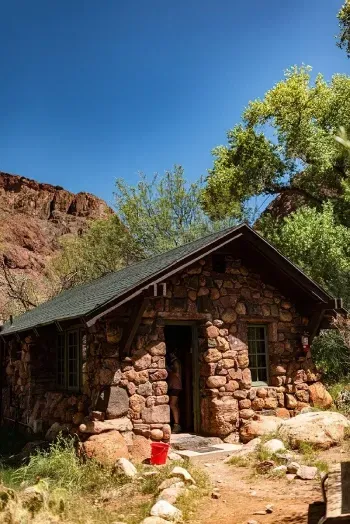 A small log cabin is surrounded by trees and rocks in the middle of a forest.