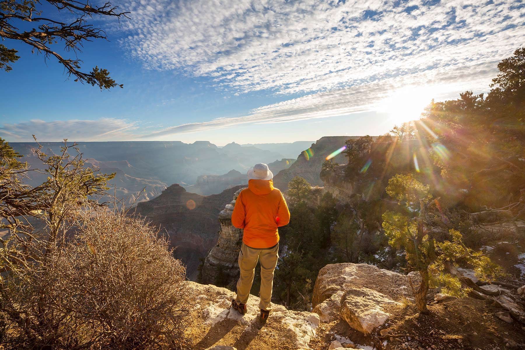 Hiker in an orange jacket stands on a rocky overlook, gazing at a sunlit mountain valley.