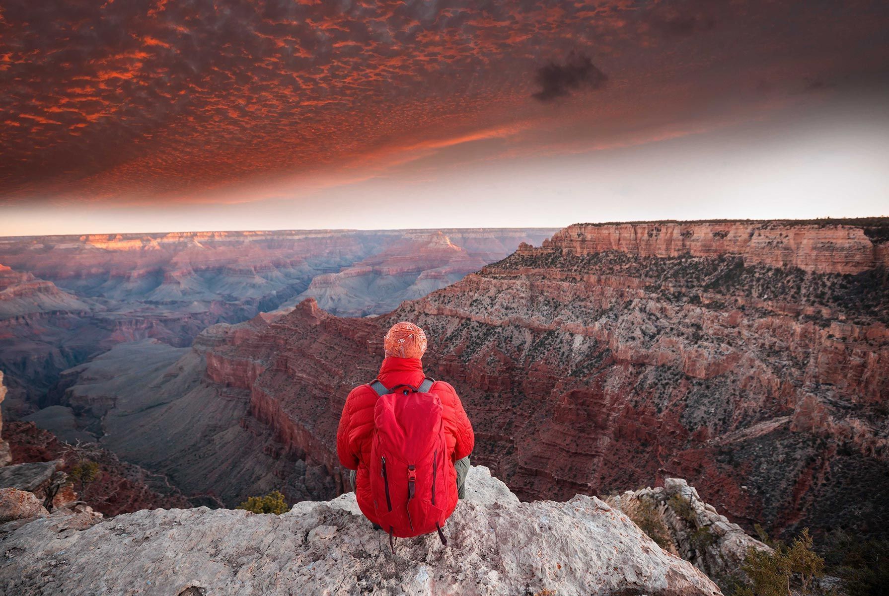 Person in a red jacket sitting on a rocky cliff edge overlooking a vast canyon at sunset