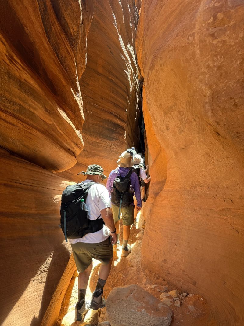 Two hikers in a narrow orange sandstone slot canyon, walking single file.