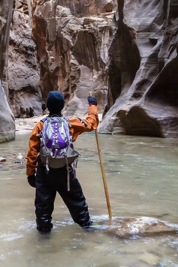 Hiker with purple backpack wades through a narrow canyon stream, holding a walking stick.