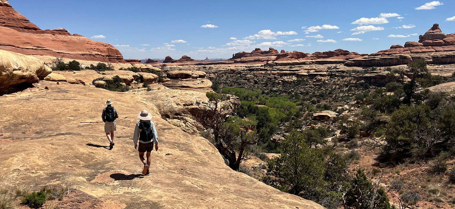 Two hikers on a desert rock trail with red cliffs and blue sky in the background