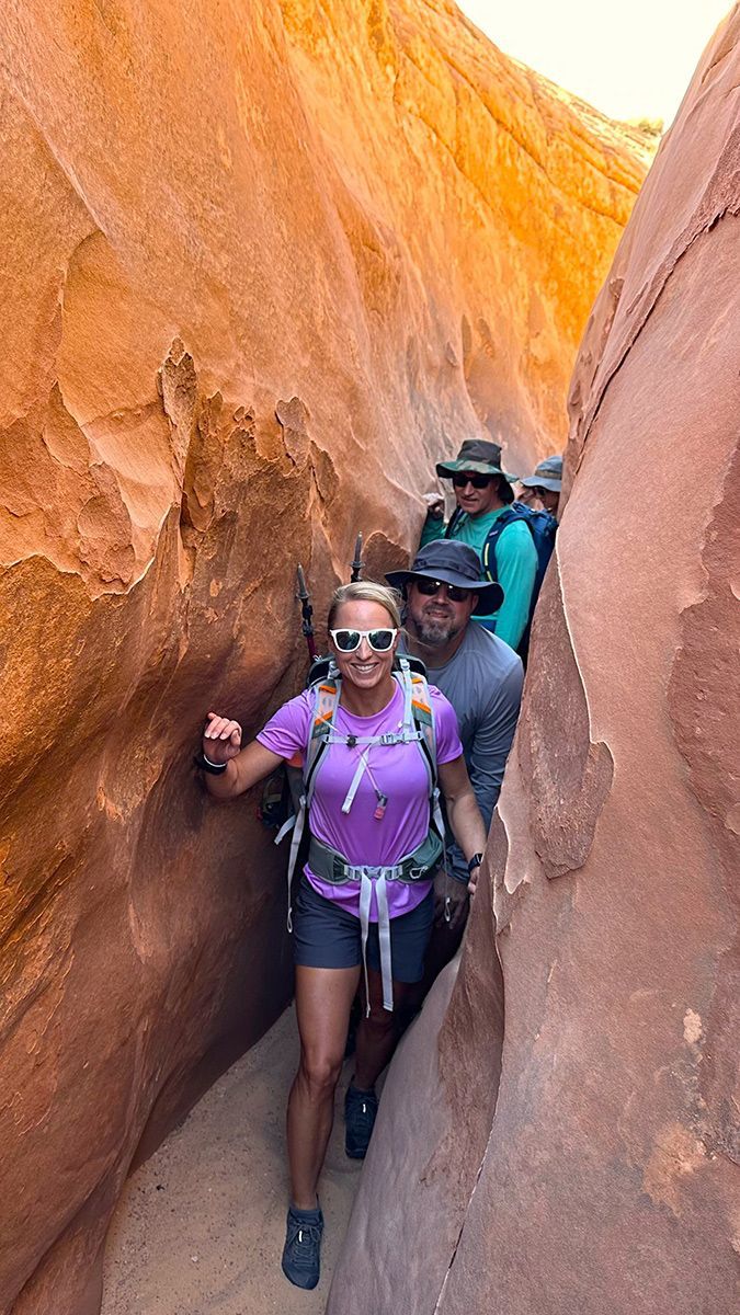 Hikers walking through a narrow orange-red canyon passage, led by a person in a purple shirt and sunglasses