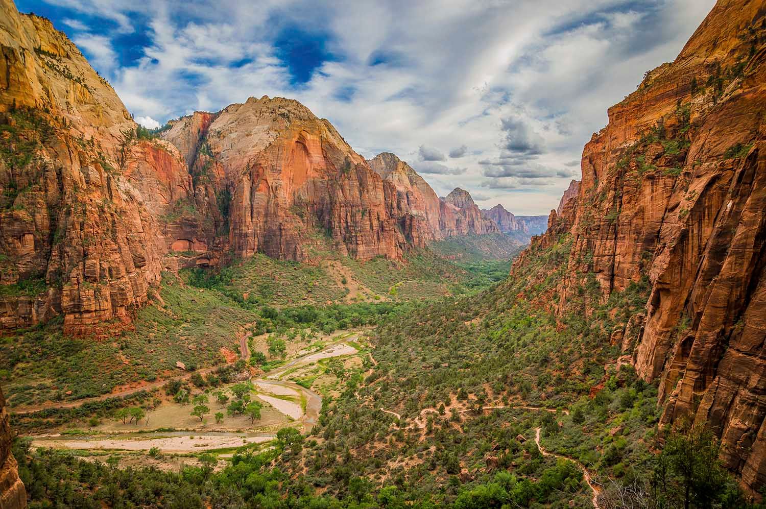 Wide canyon with red sandstone cliffs, a green valley, and a winding river under a cloudy sky