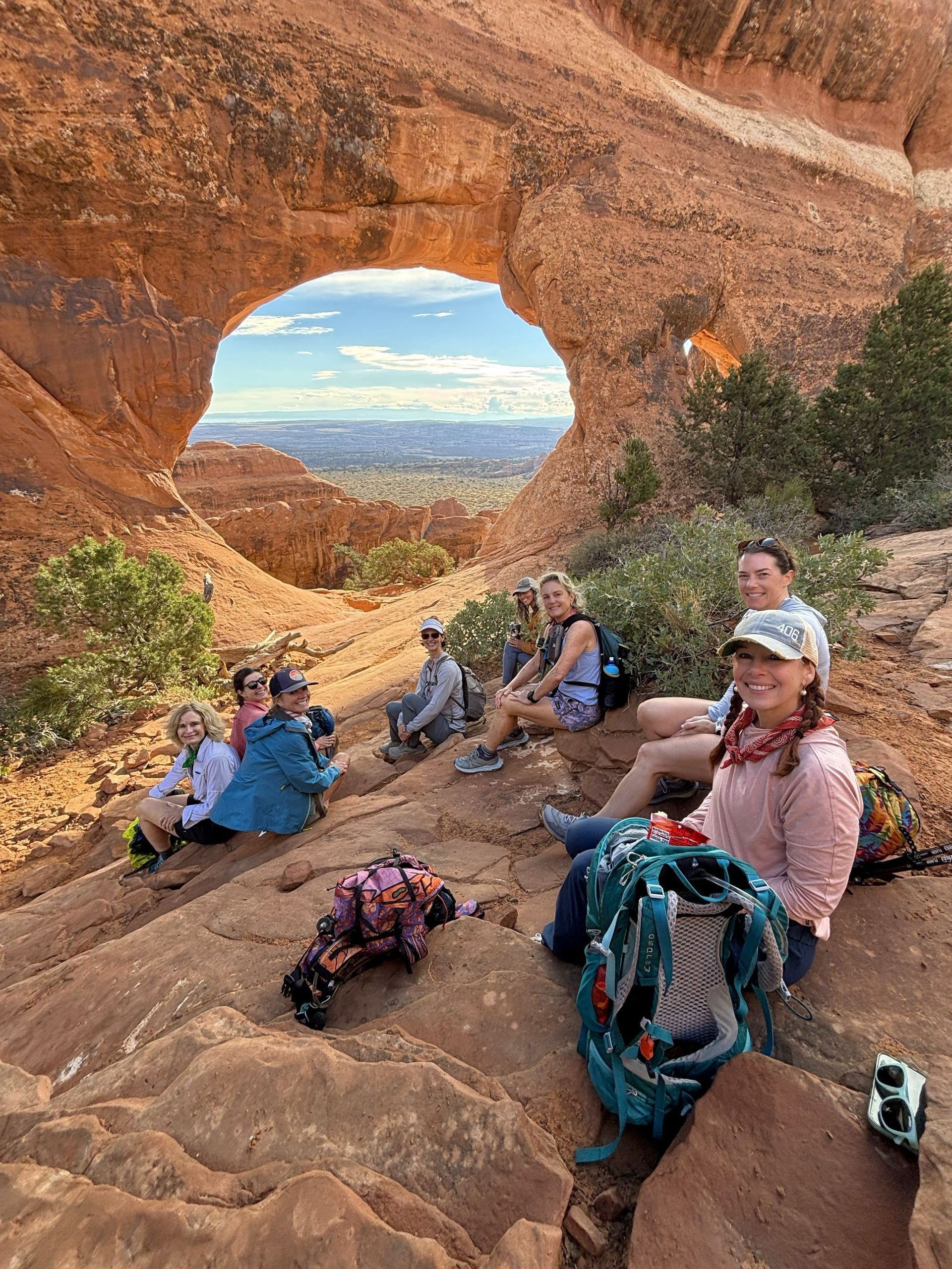 Hikers resting on a sandstone ledge inside a rock arch, overlooking a desert valley at sunset.
