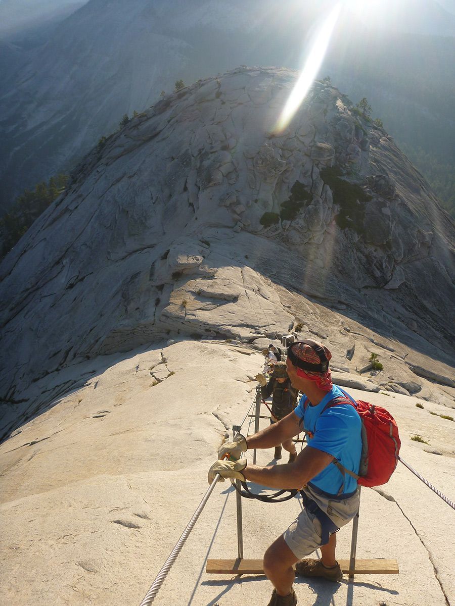 Climber in blue shirt and red backpack rappels down a steep rocky ridge in bright sunlight.