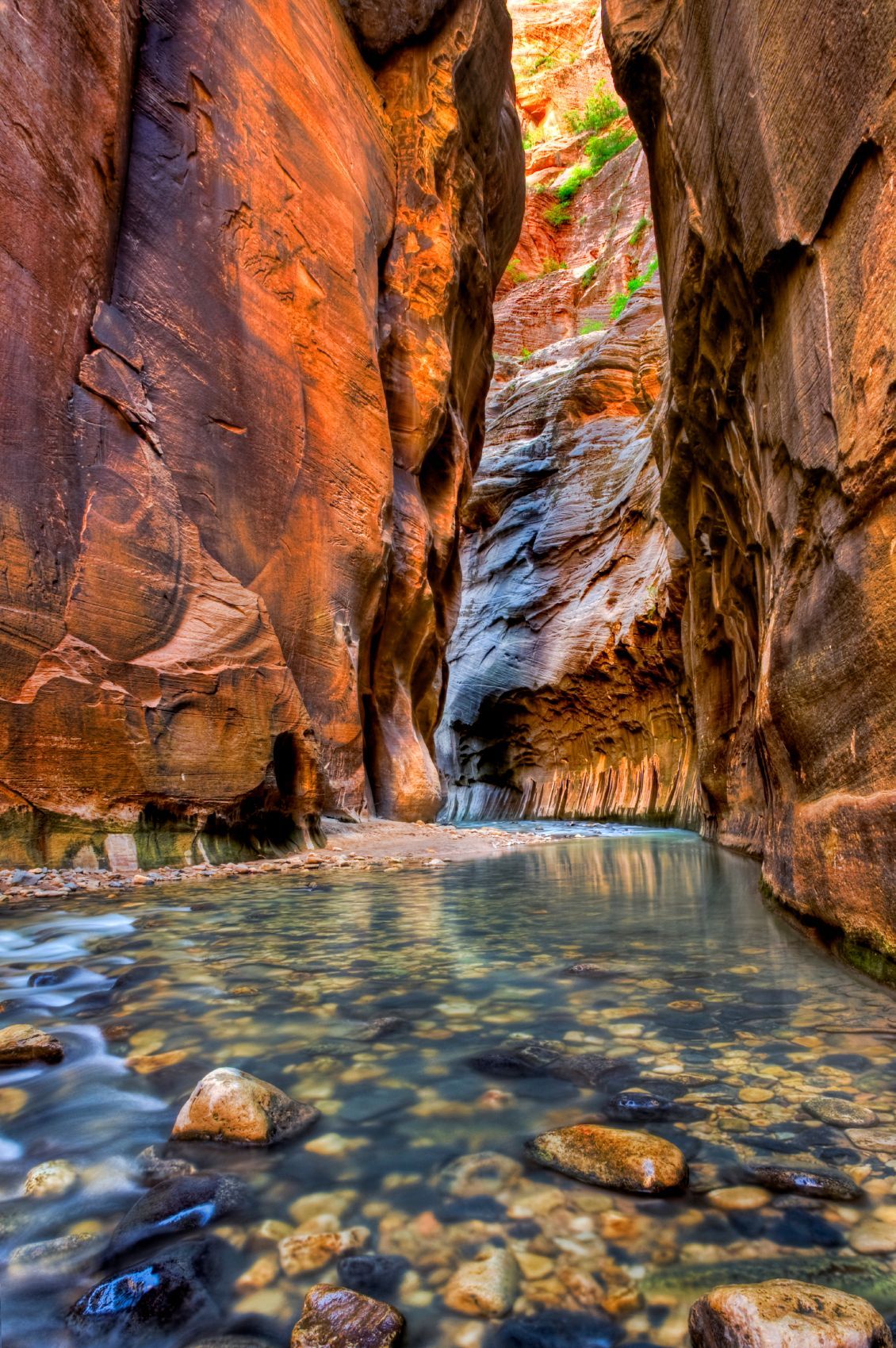 Narrow canyon with orange rock walls and a clear shallow stream over pebbles