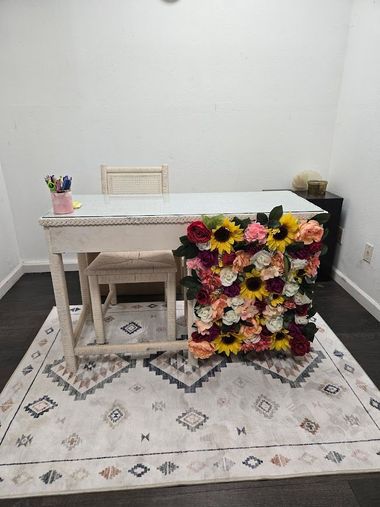White desk and chair in a sparsely decorated room, with a large colorful floral arrangement and patterned rug.
