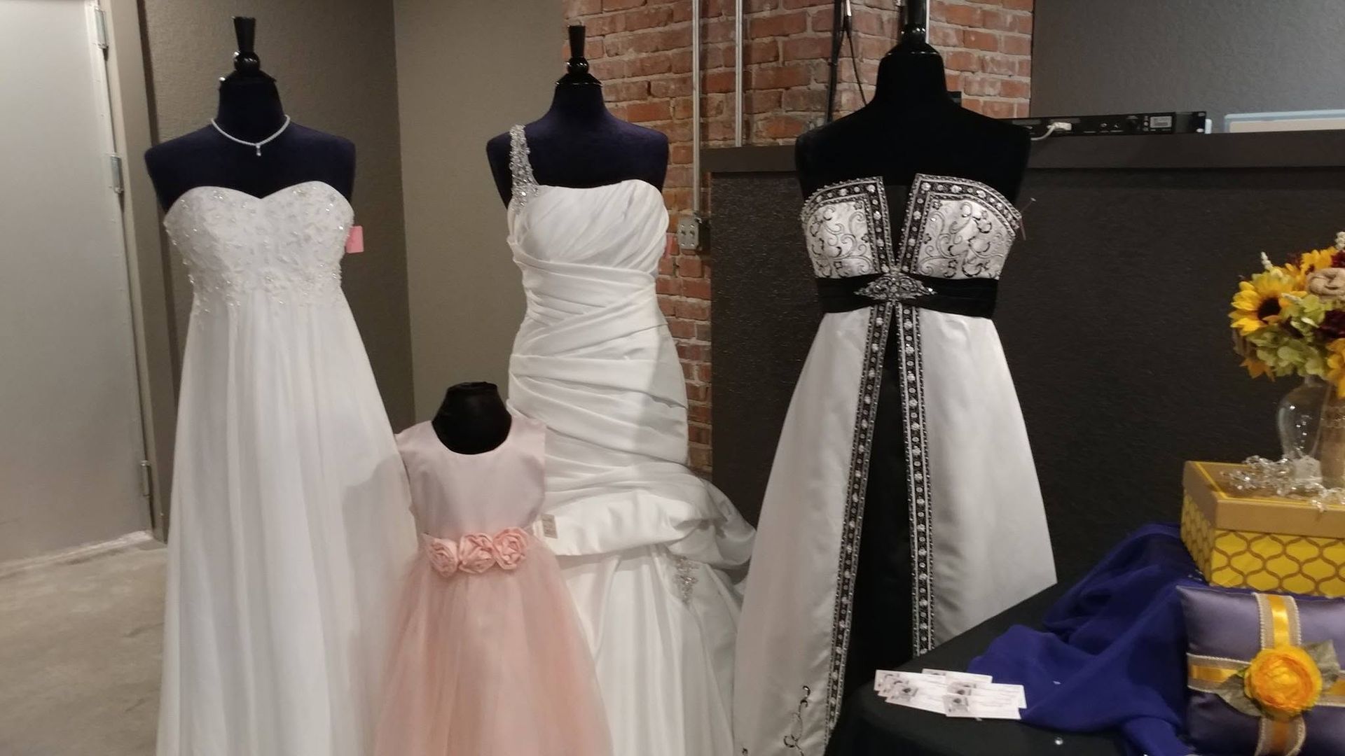 Three mannequins display white and black formal dresses in a boutique, with a pink child dress in front.