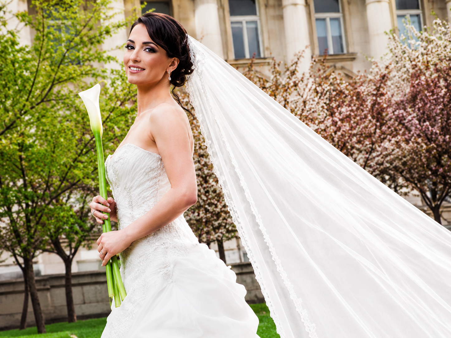 Bride in a white gown holding a bouquet outdoors, with a long veil flowing behind her.