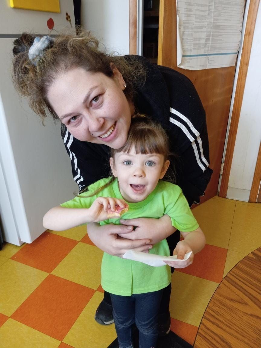 An adult stands behind and hugs a child in a green shirt, both smiling in a room with a yellow and orange tiled floor.