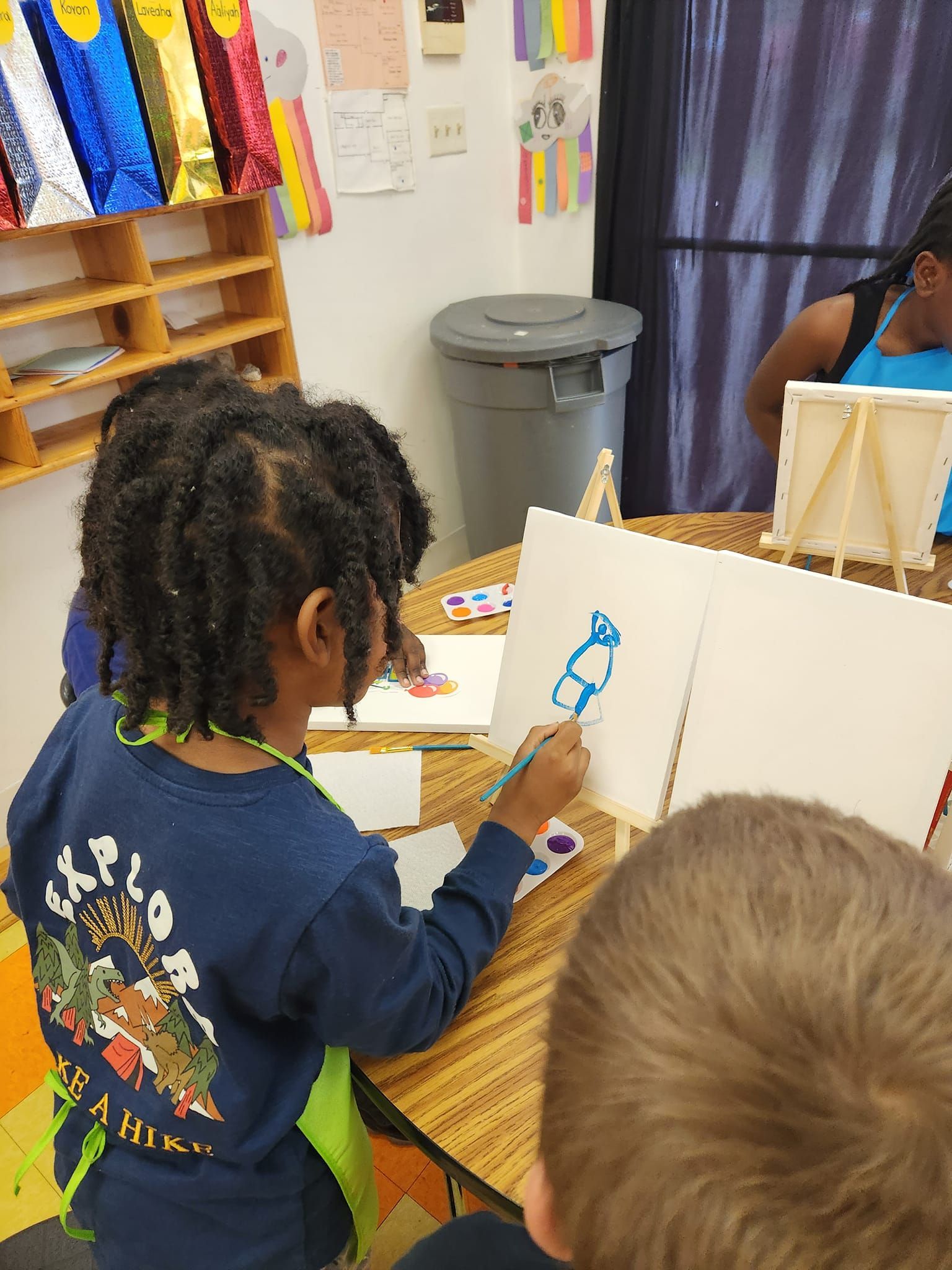 A child with dark braids wearing a blue long-sleeve shirt paints a blue shape on a canvas at a table in a bright room.