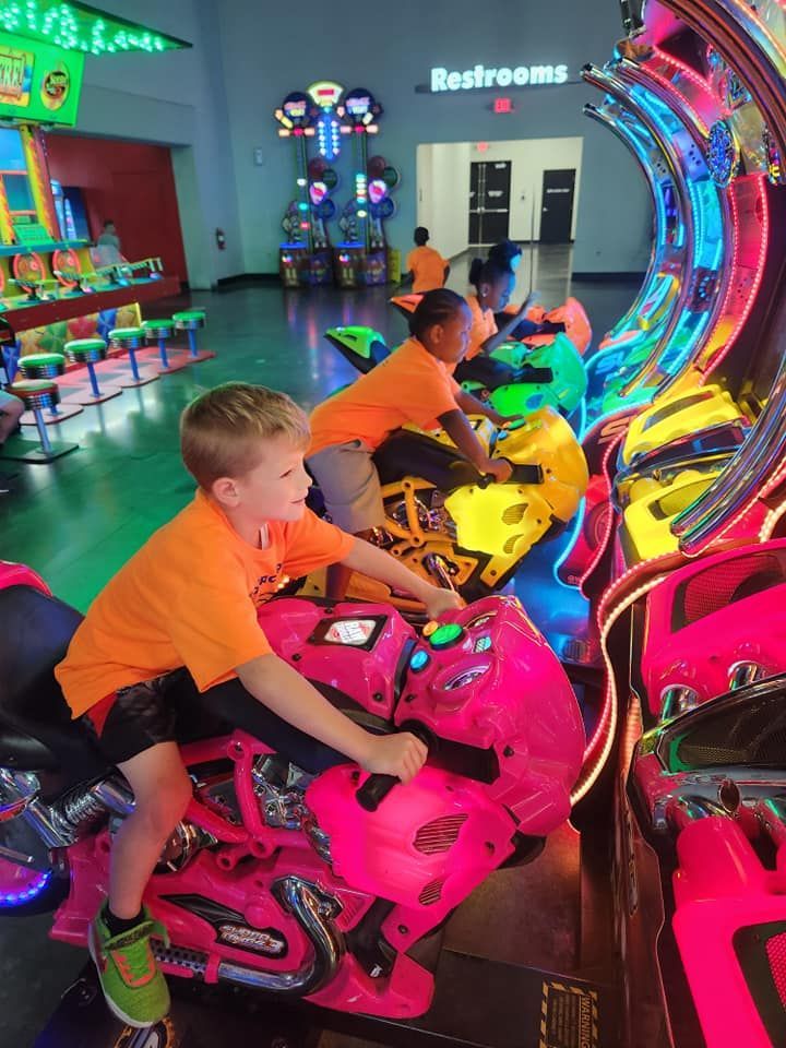 Children in bright orange shirts play on bright pink and yellow motorcycle arcade games in an indoor entertainment center.