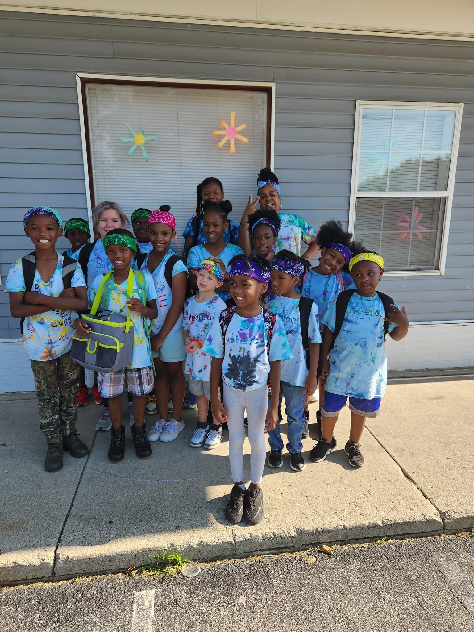 A group of children wearing matching blue tie-dye shirts and headbands stand outdoors in front of a building.