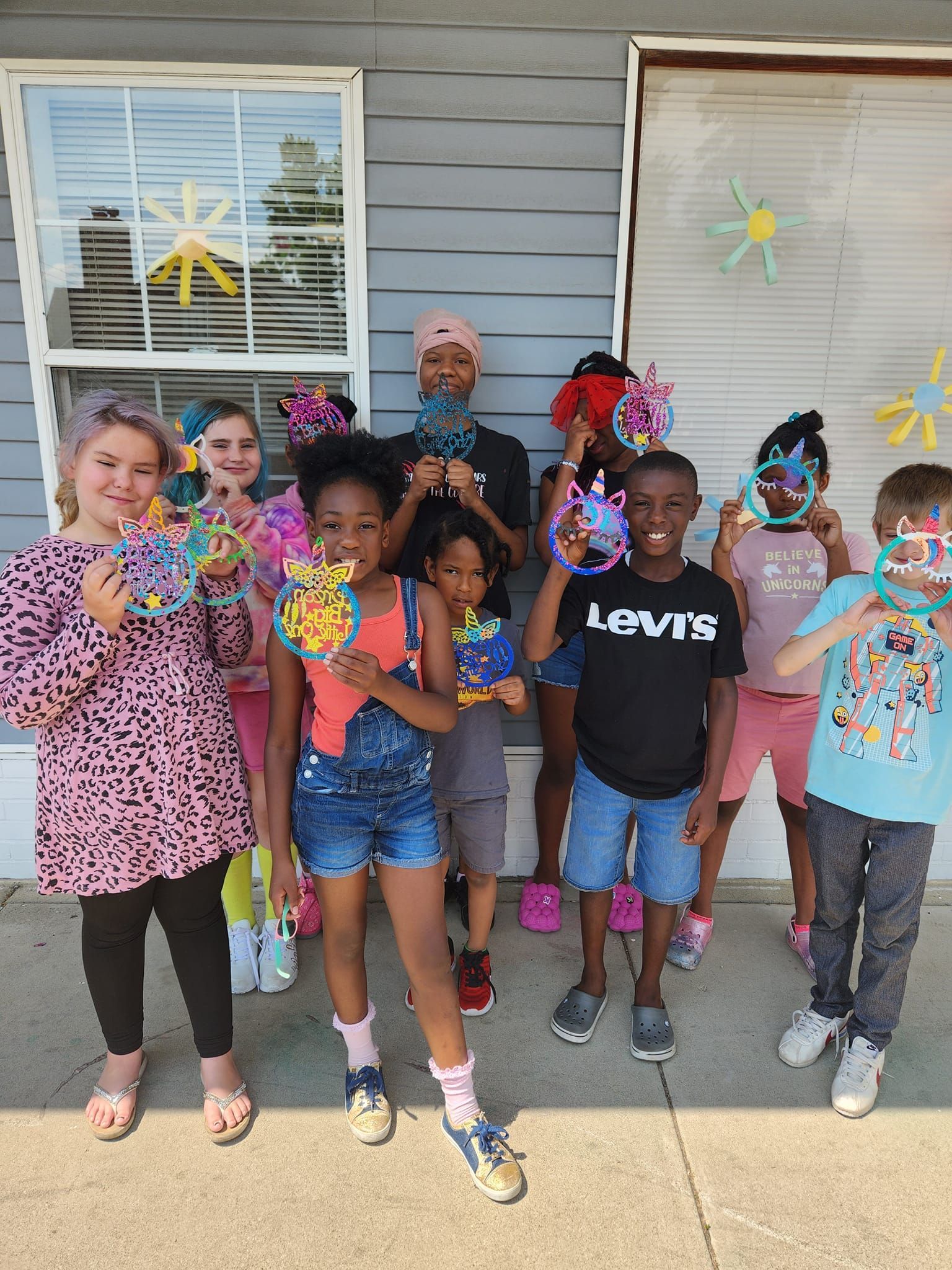 A group of children stand outside a gray-sided building, holding colorful paper crafts in front of their faces.