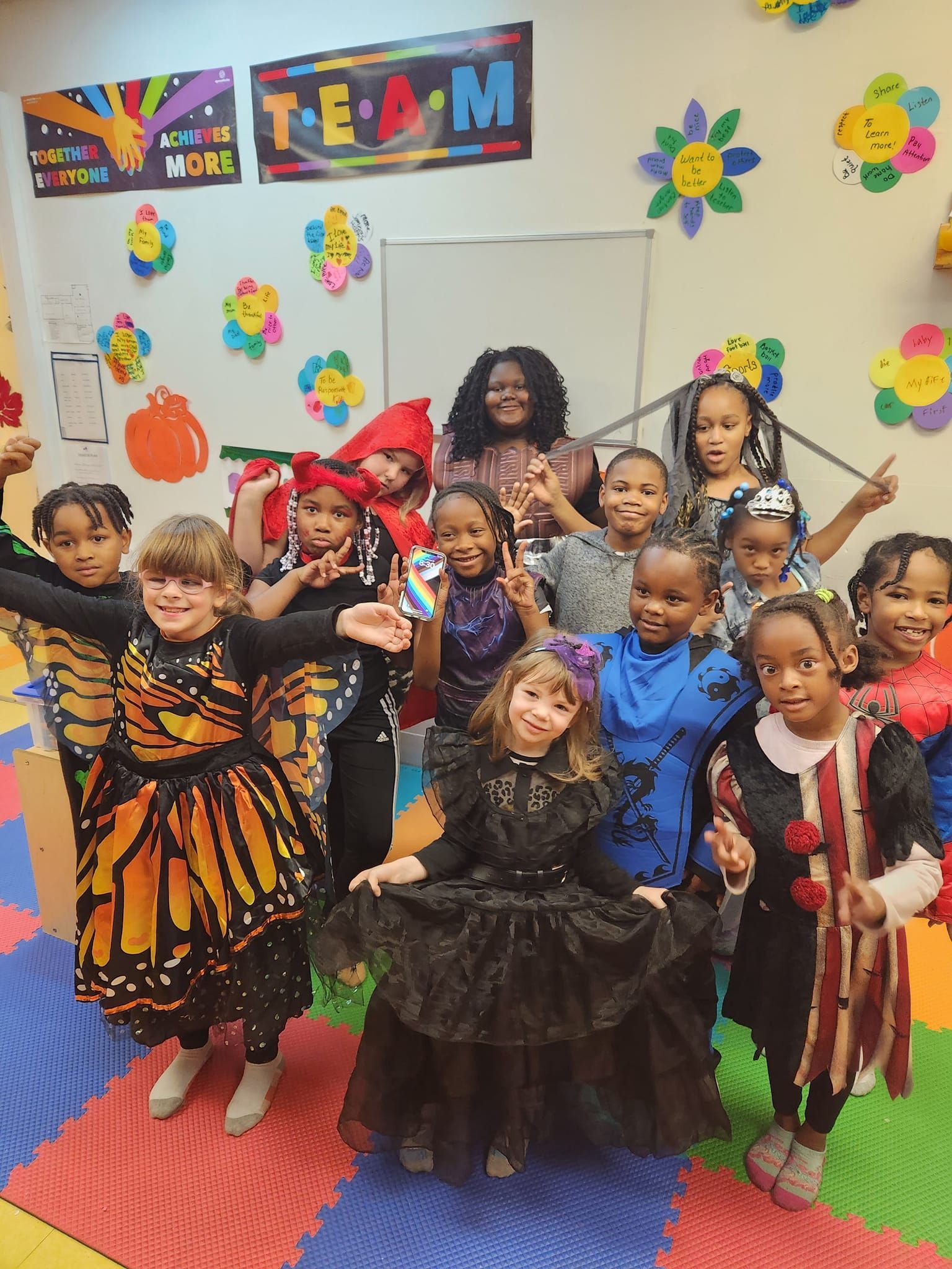 A group of children and an adult in Halloween costumes stand together in a colorful, decorated classroom.