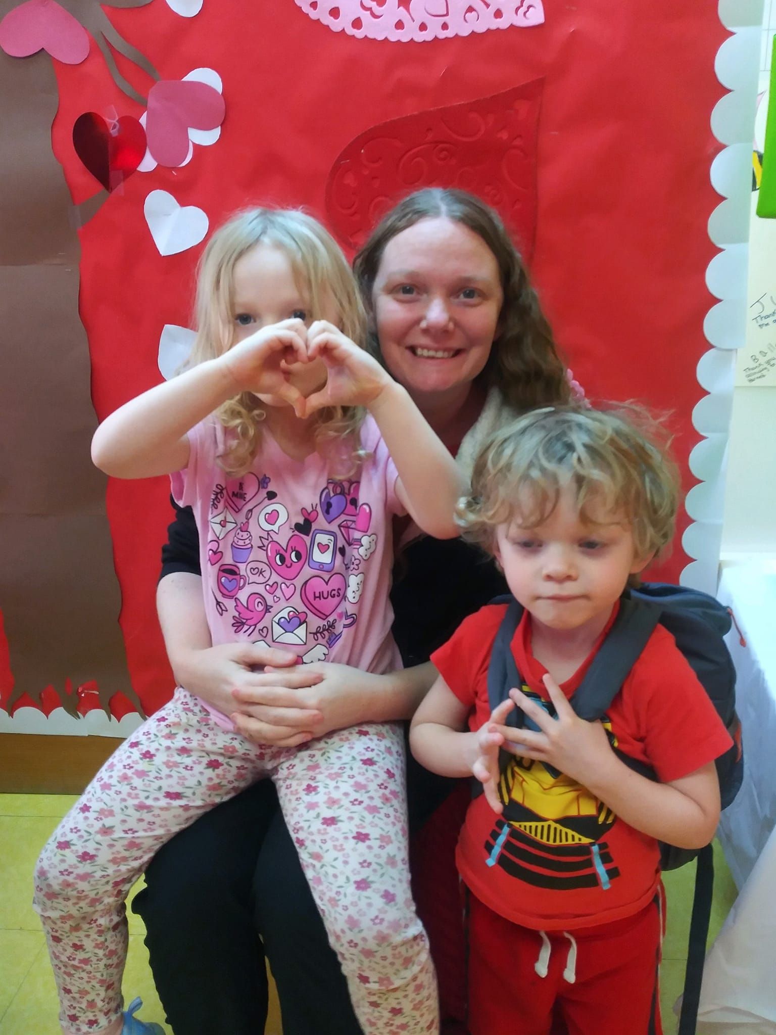 A person sits with two children in front of a red heart-themed display; one child makes a heart shape with their hands.