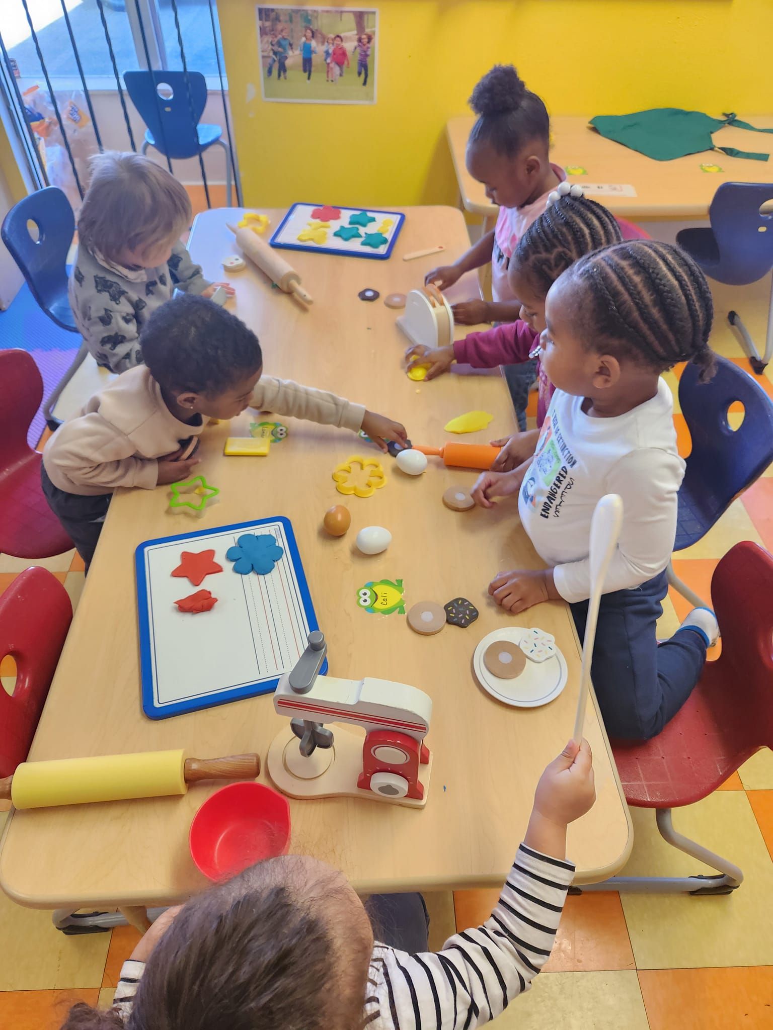Children play with colorful toy kitchen items and shapes at a table in a classroom setting.