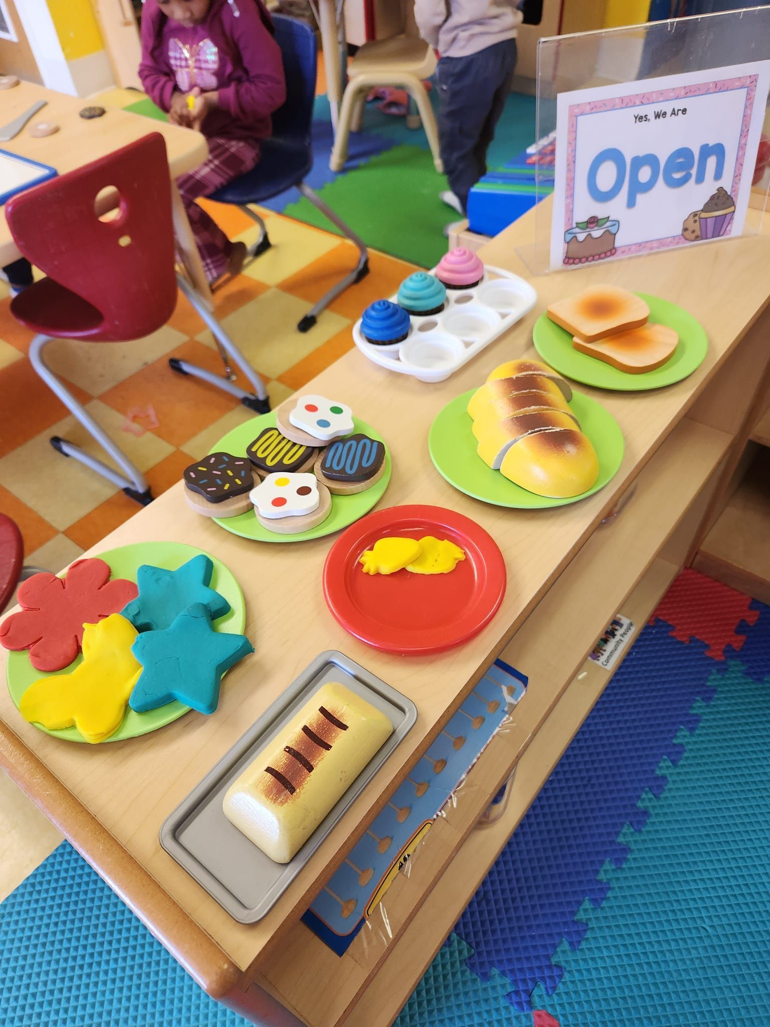 A bakery-themed play station with toy food on plates, including pastries, bread, and cookies, next to an 