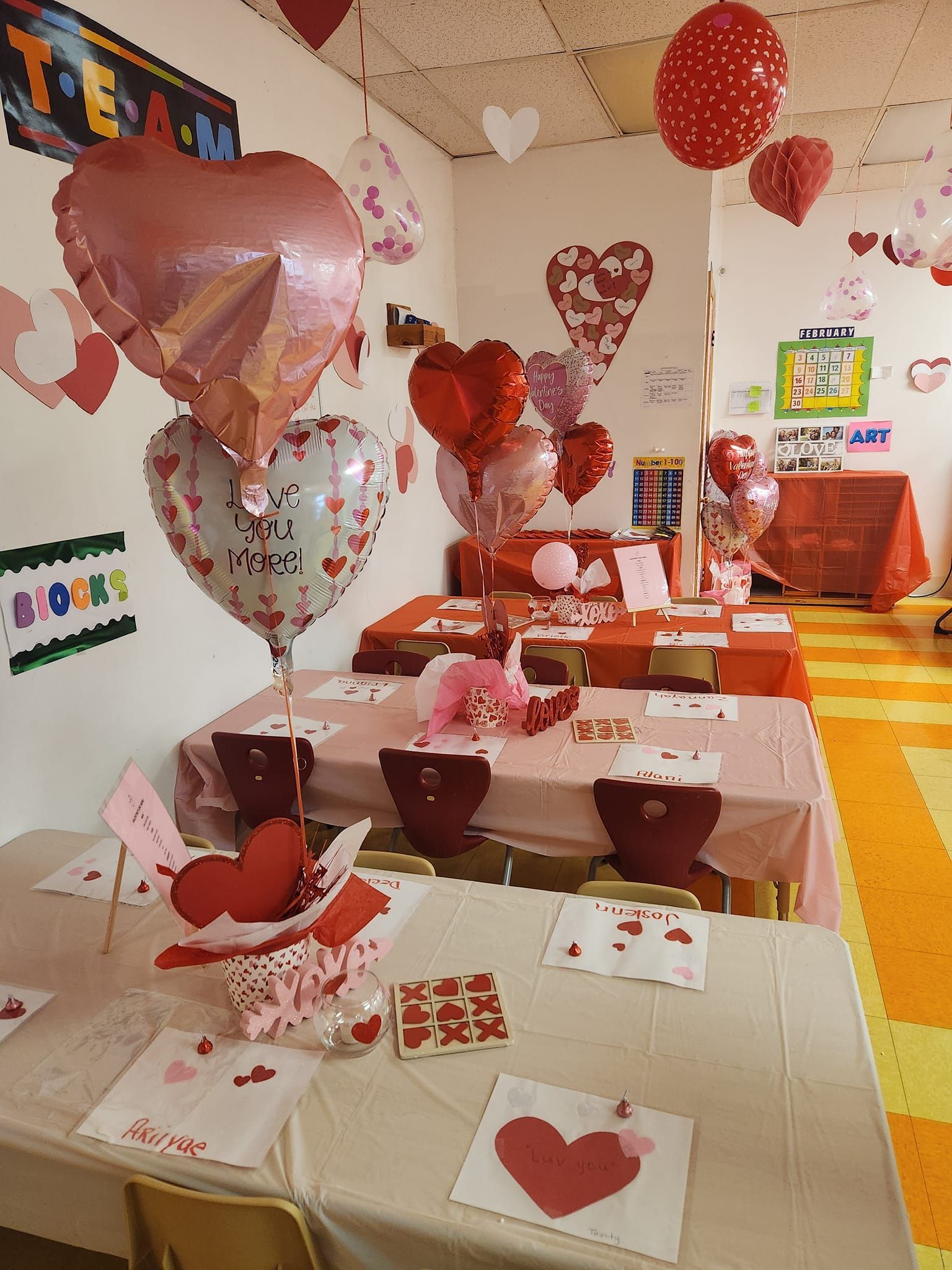 A room decorated for Valentine's Day with heart-shaped balloons, pink tablecloths, and heart-themed activities on tables.