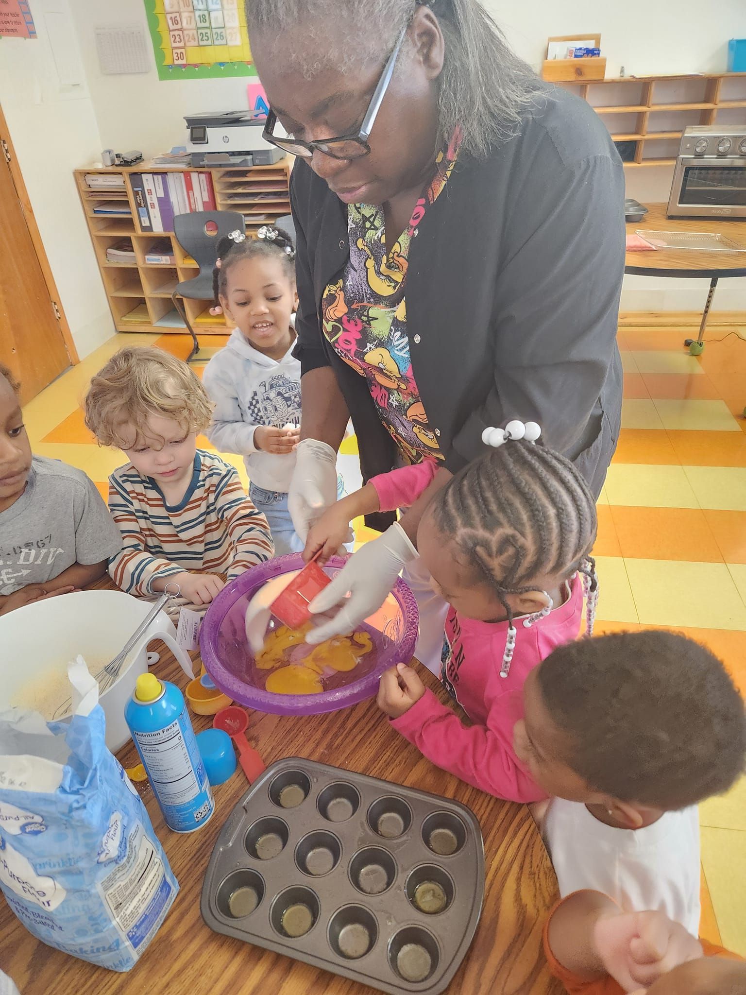 A teacher and four students prepare muffin batter in a purple bowl in a brightly lit classroom.