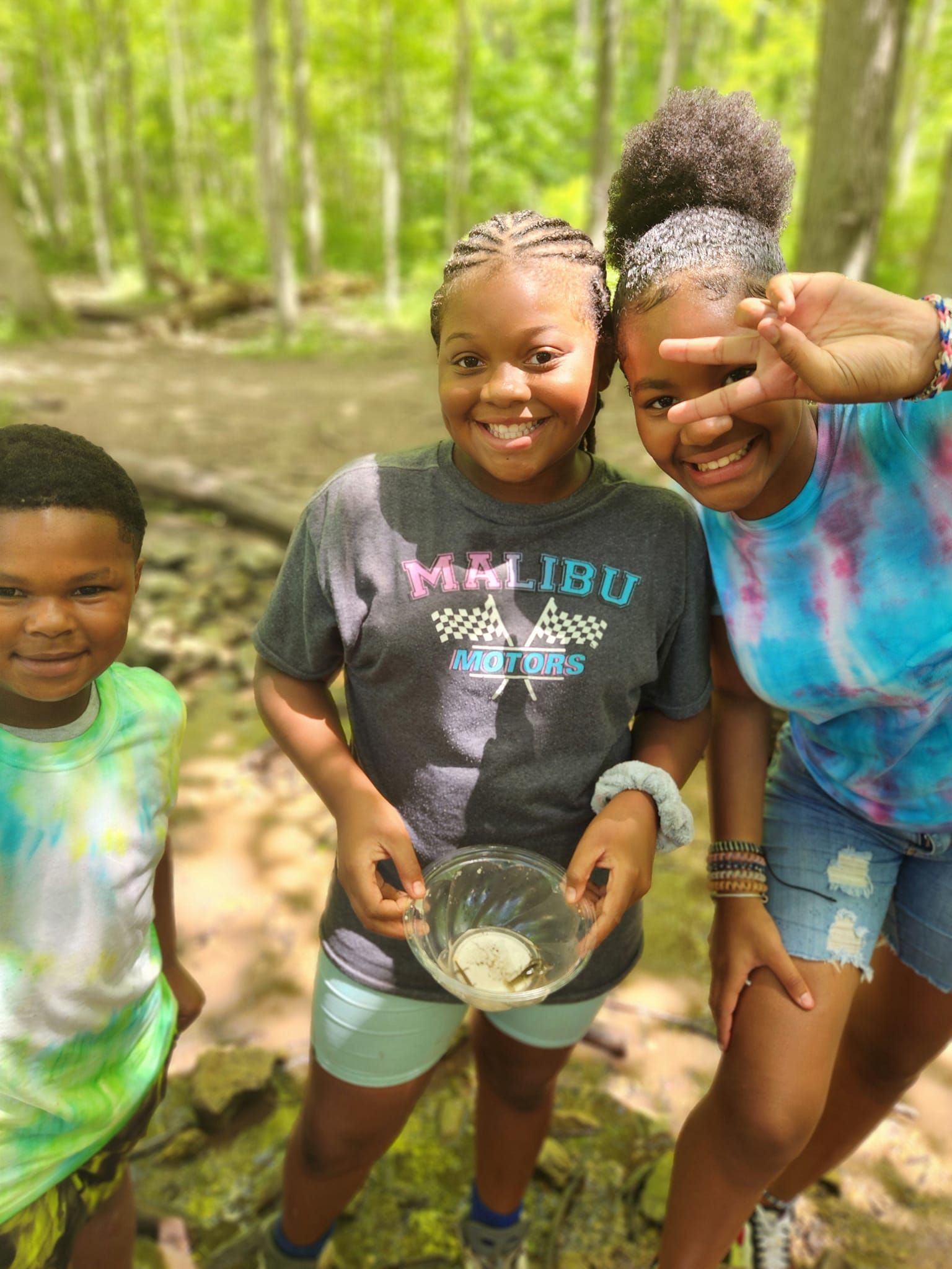 Three children smile outdoors in a forest. The center child holds a small, clear bowl. One makes a peace sign gesture.