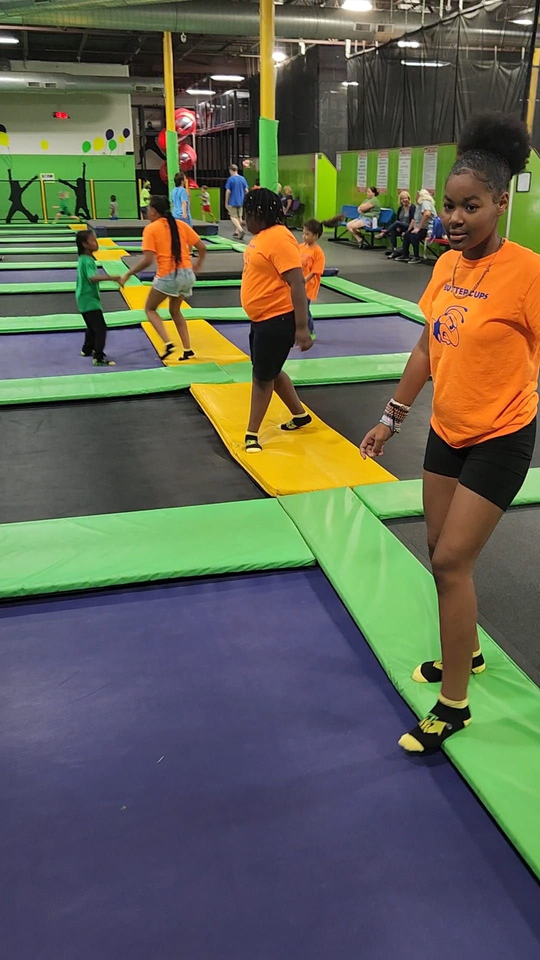 People in bright orange t-shirts jumping on interconnected trampolines at an indoor recreation center.