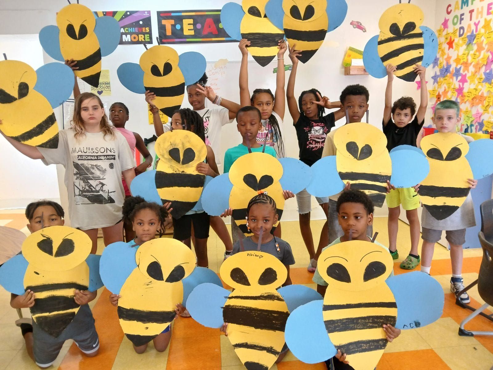 A group of children hold large, handmade paper bumblebee crafts in a colorful classroom.