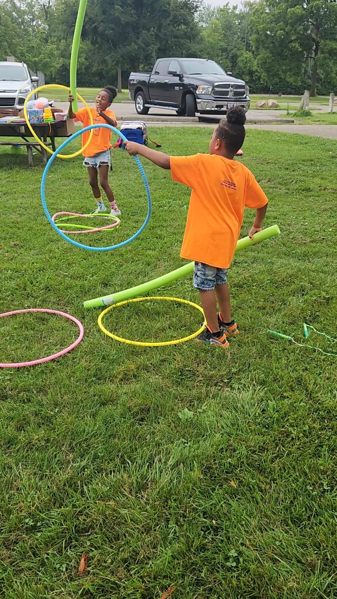 Two children in bright orange shirts play with colorful hula hoops and pool noodles in a grassy park.
