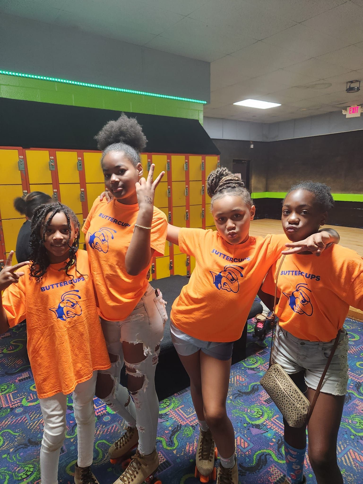 Four people in matching orange shirts and roller skates stand together in front of a wall of yellow lockers.