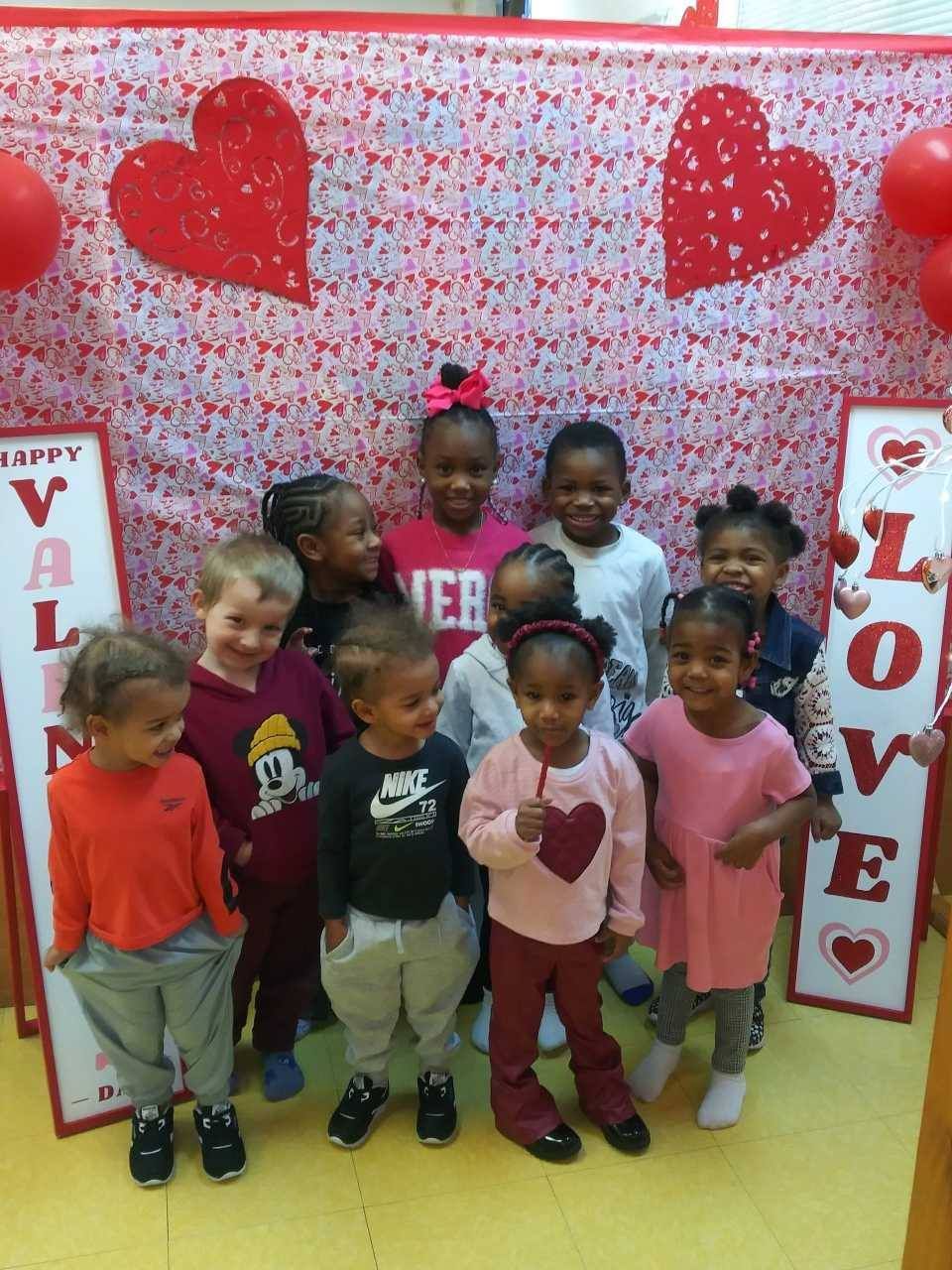 A group of children posing for a photo in front of a Valentine's Day backdrop with red hearts and 