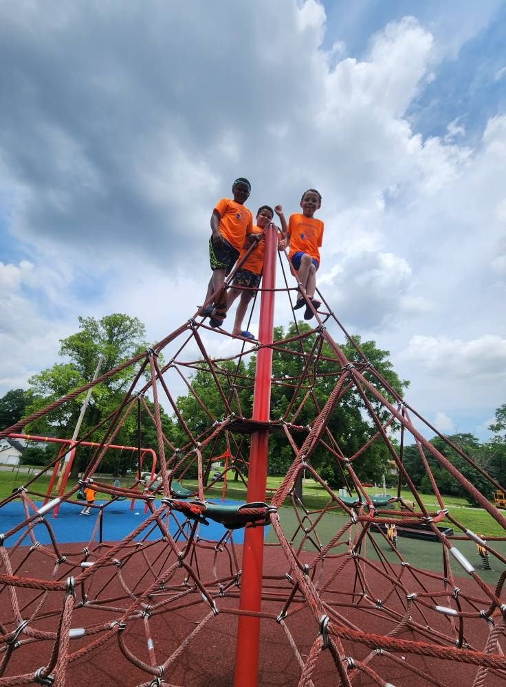 Three children in bright orange shirts climb to the top of a red pyramid-shaped rope structure at a playground.