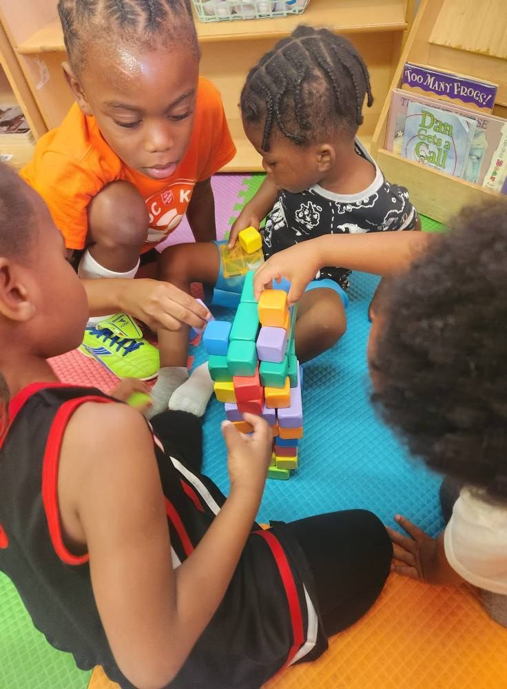 Children sit on a colorful foam mat, working together to build a tall, multi-colored tower out of plastic blocks.