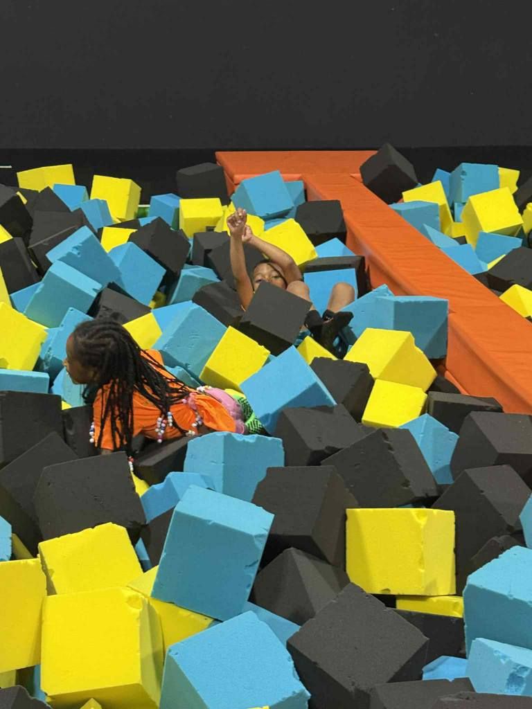 Two people play in a foam pit filled with blue, yellow, and black cubes next to an orange padded platform.