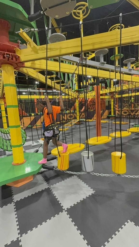 A person in an orange shirt and safety harness navigates an indoor obstacle course with hanging yellow platforms.