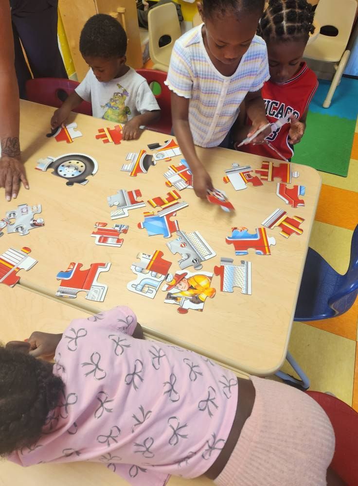 Four children sit at a table in a classroom working together on a colorful floor puzzle.