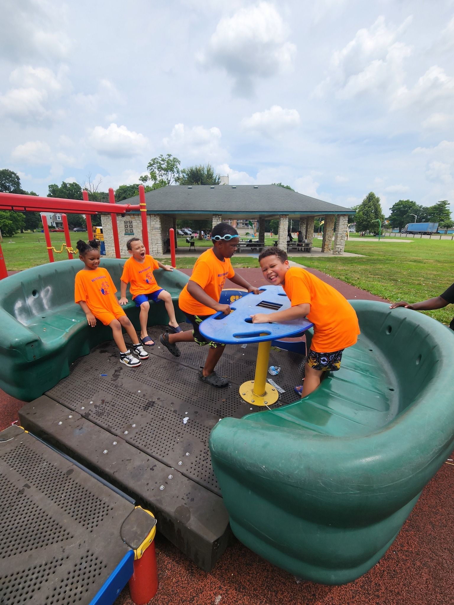 Four children in matching orange shirts play on a green and blue circular playground merry-go-round in a park.