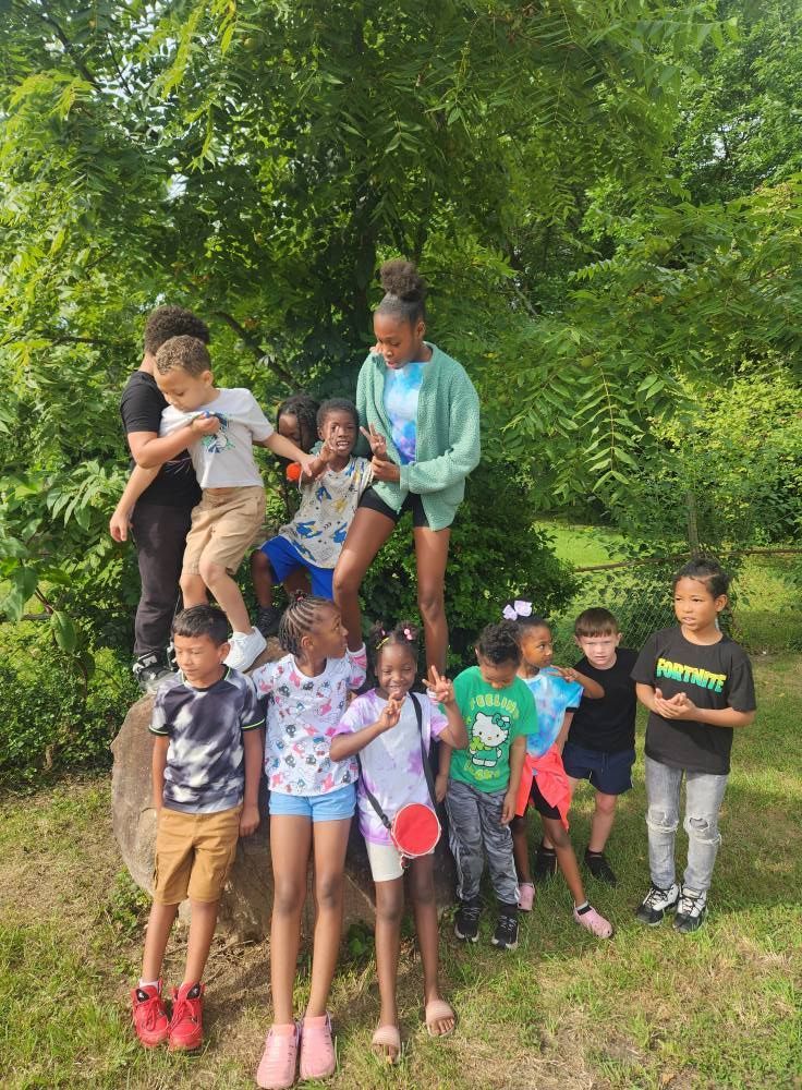 A group of children stand and climb on a large tree stump in a lush, green outdoor setting.