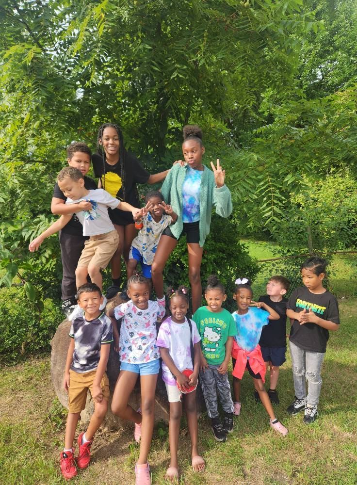 A group of children and two young adults pose together outdoors on a large tree stump and on the grass.