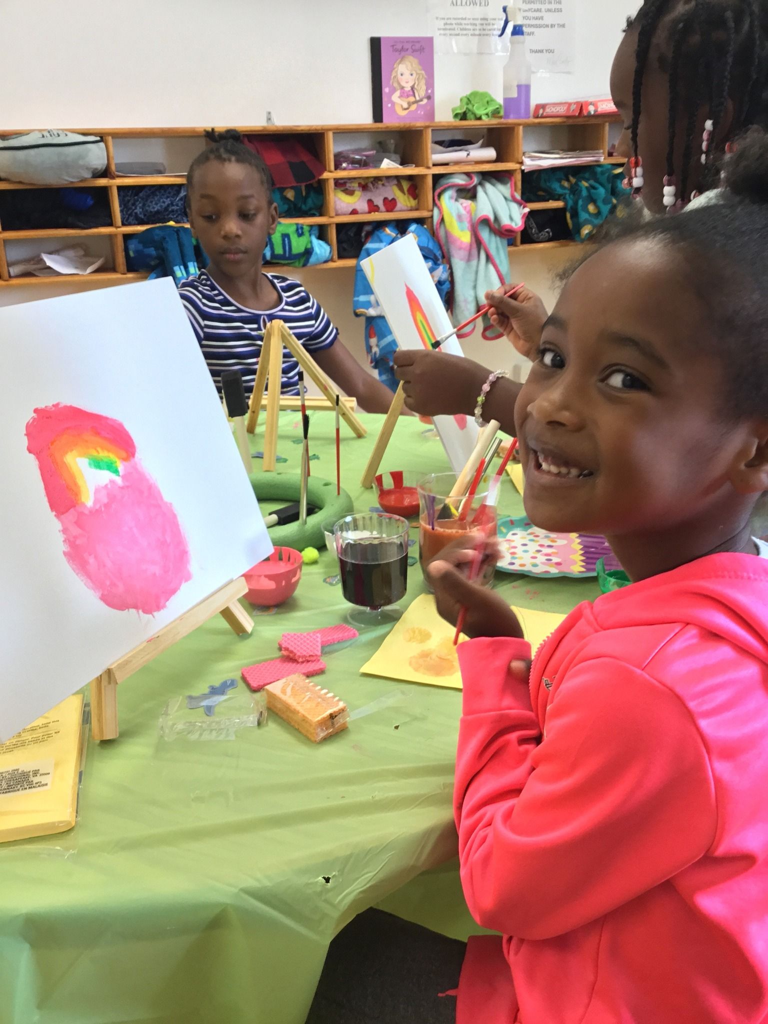 Two children painting on canvases at a green-covered table in a classroom setting.