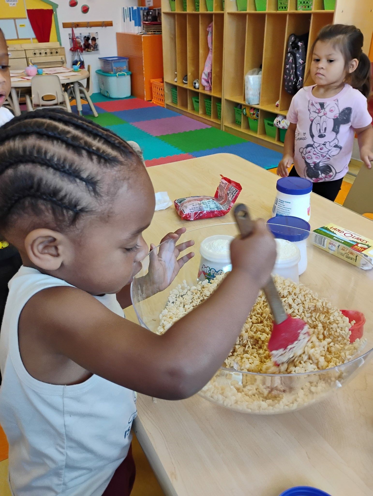 A young child mixes ingredients in a bowl on a classroom table, while another child observes in the background.