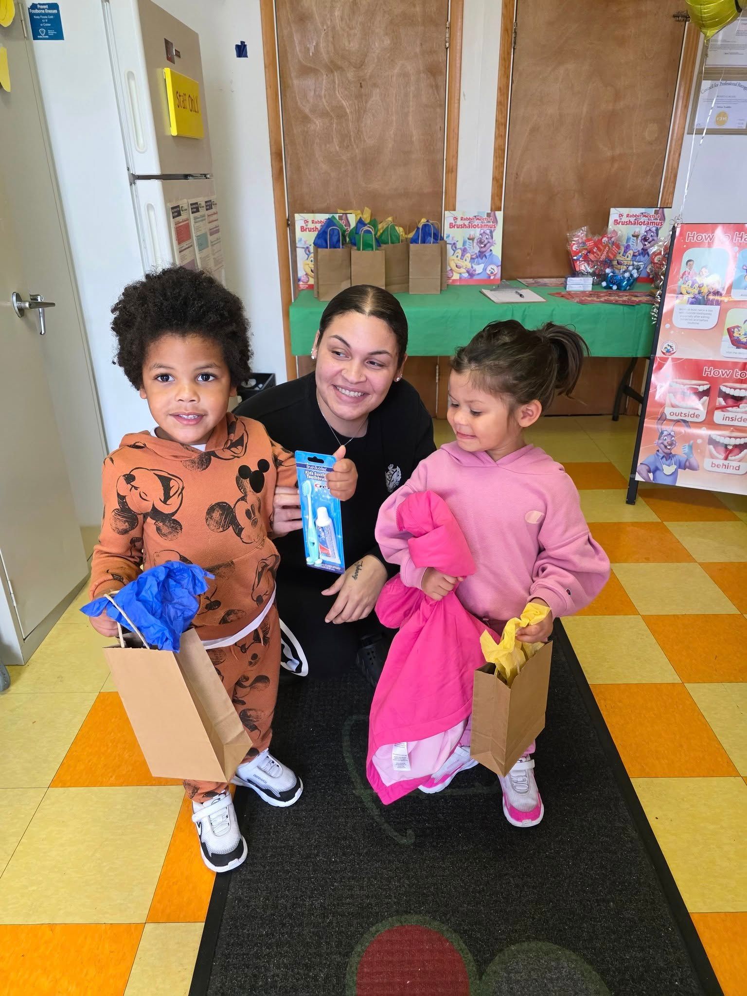 A smiling person holds gift bags for two children in a room with a green table and yellow and white checkered flooring.