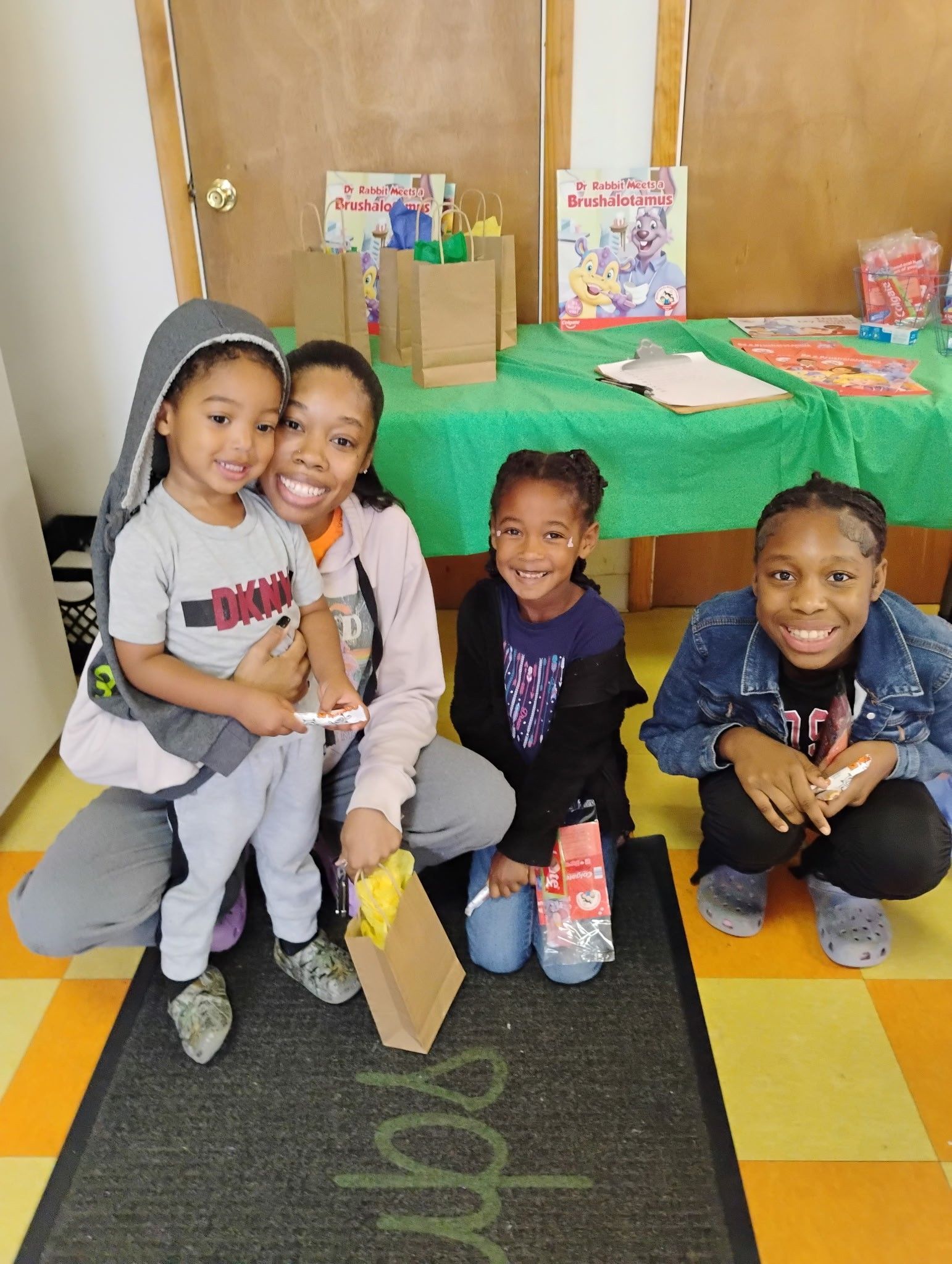 A smiling adult and three children pose in front of a green-covered table with bags and books in a bright indoor room.