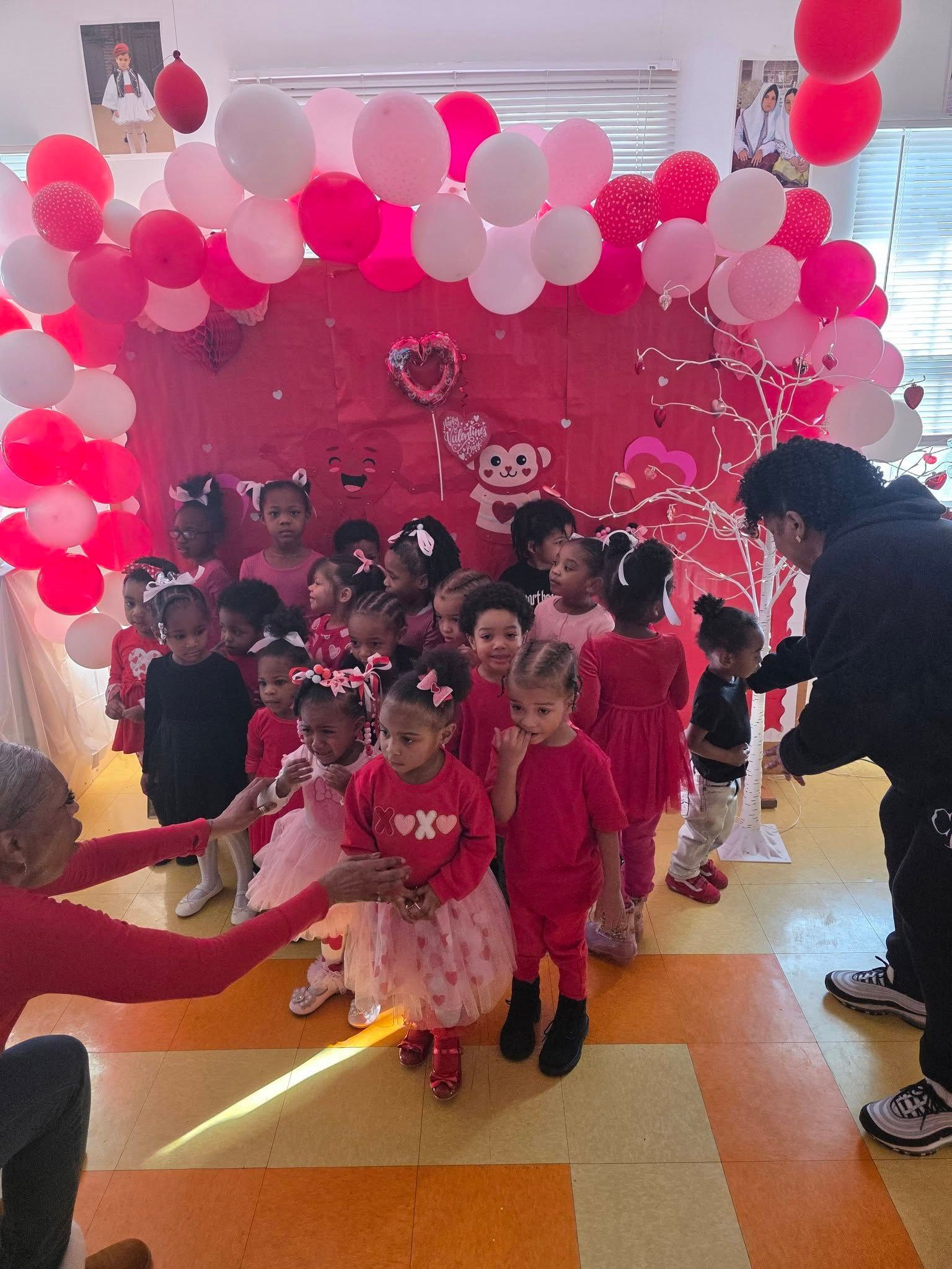 A group of children dressed in red and pink standing in front of a red, balloon-decorated backdrop for a party.