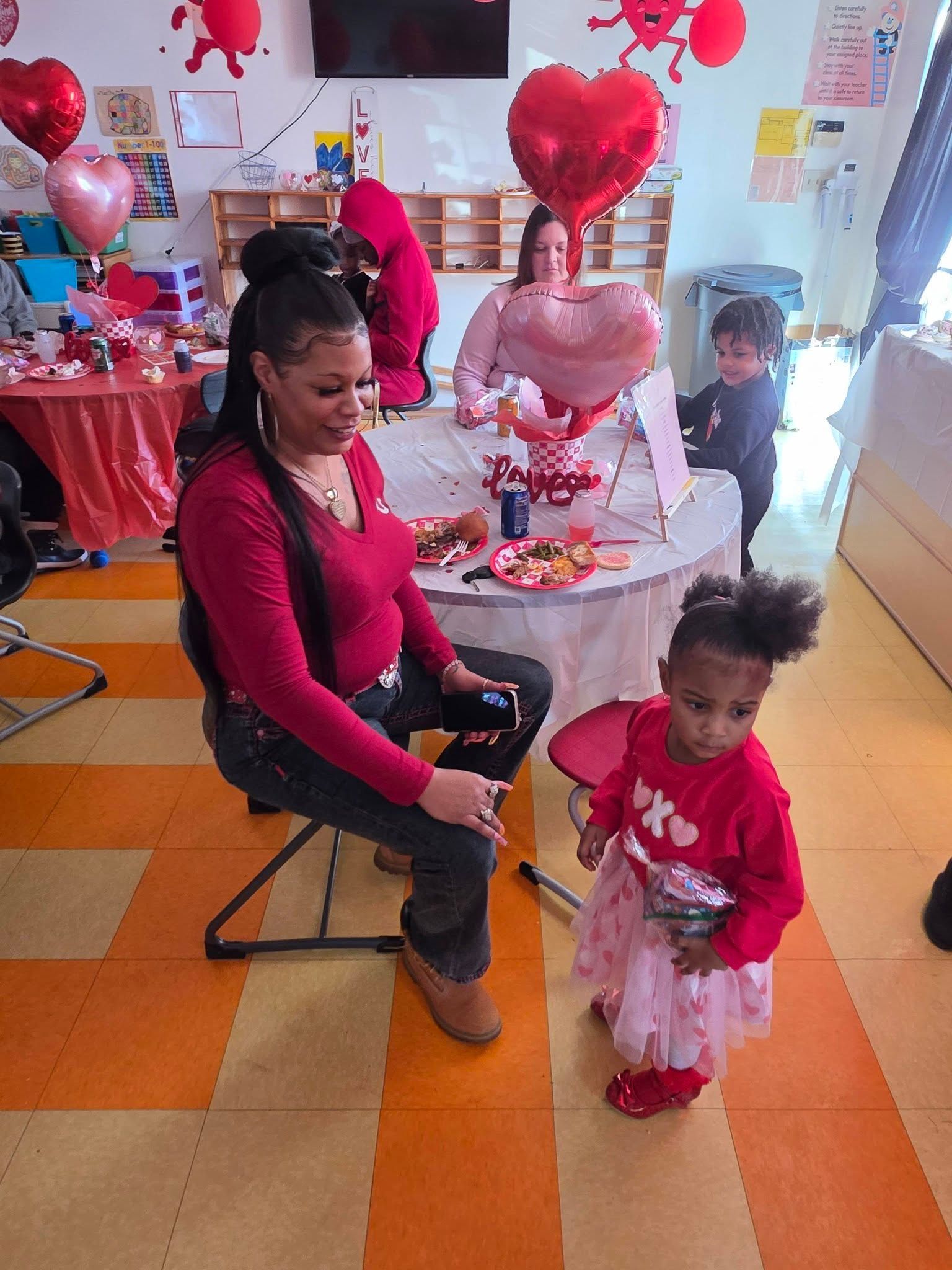 A woman in a red top and a child in a pink skirt pose in a decorated room with heart balloons for a Valentine's event.