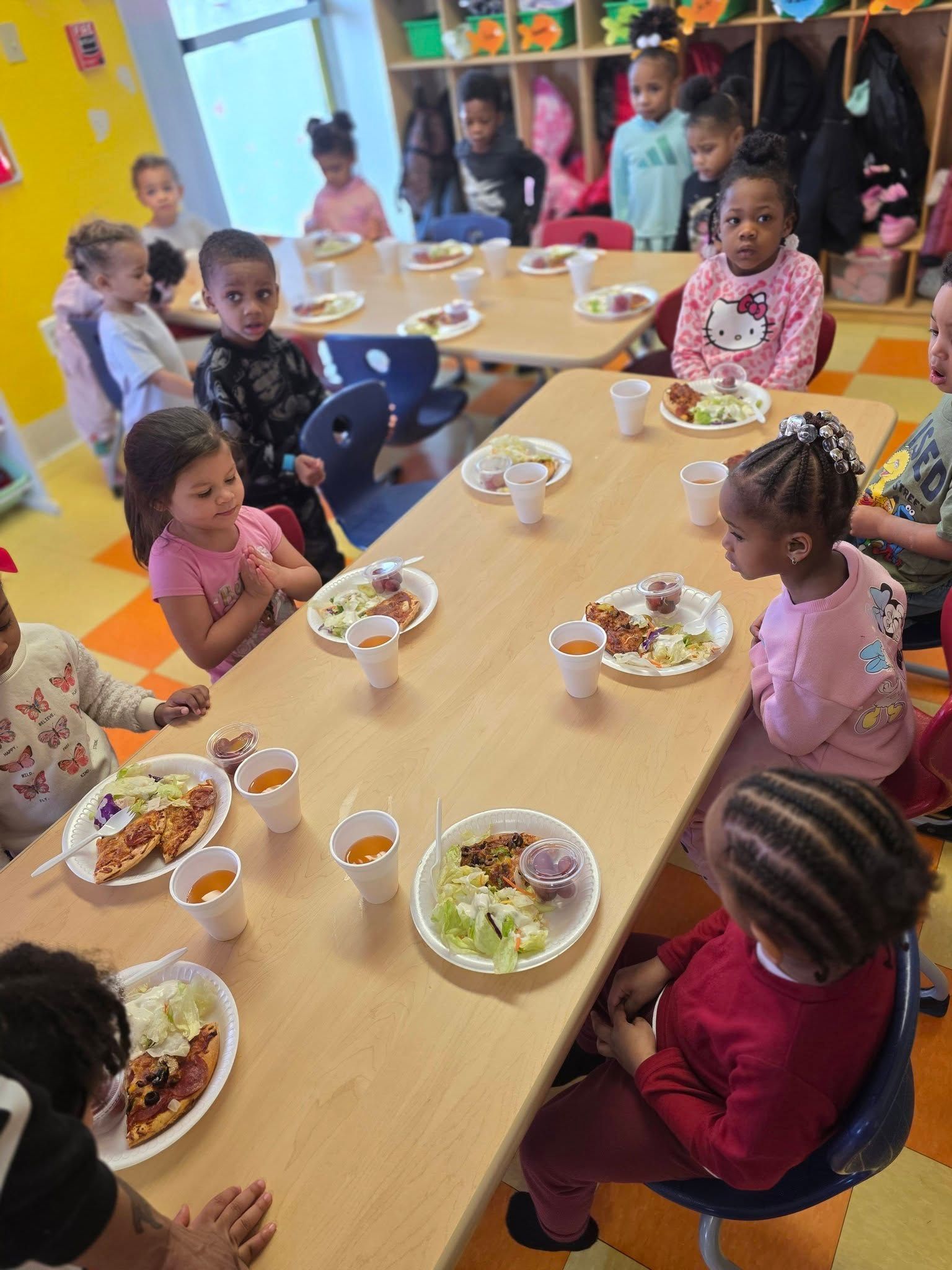 Children sit at tables in a bright classroom, eating lunch from plates with portions of food and cups on a light wood table.
