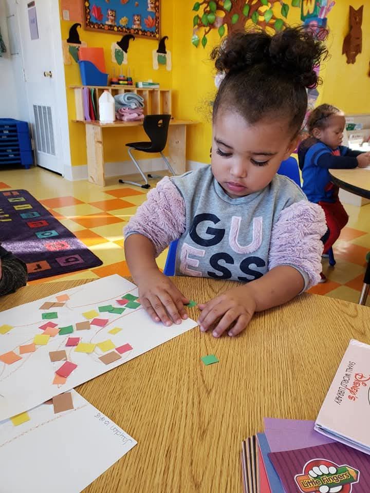A child works on a colorful paper collage at a wooden table in a classroom with yellow walls.
