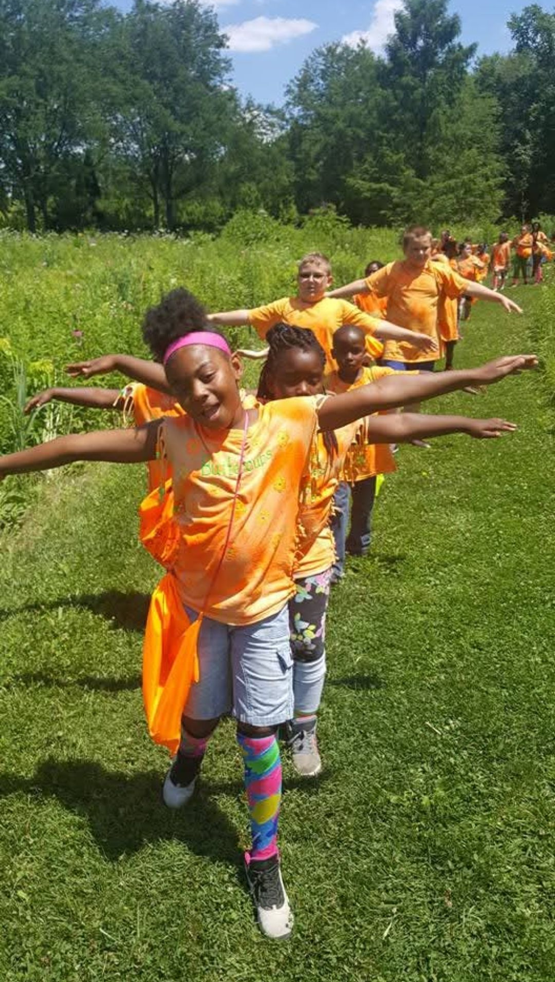 A group of people wearing matching orange shirts stand in a line on a grassy field, holding their arms out to their sides.