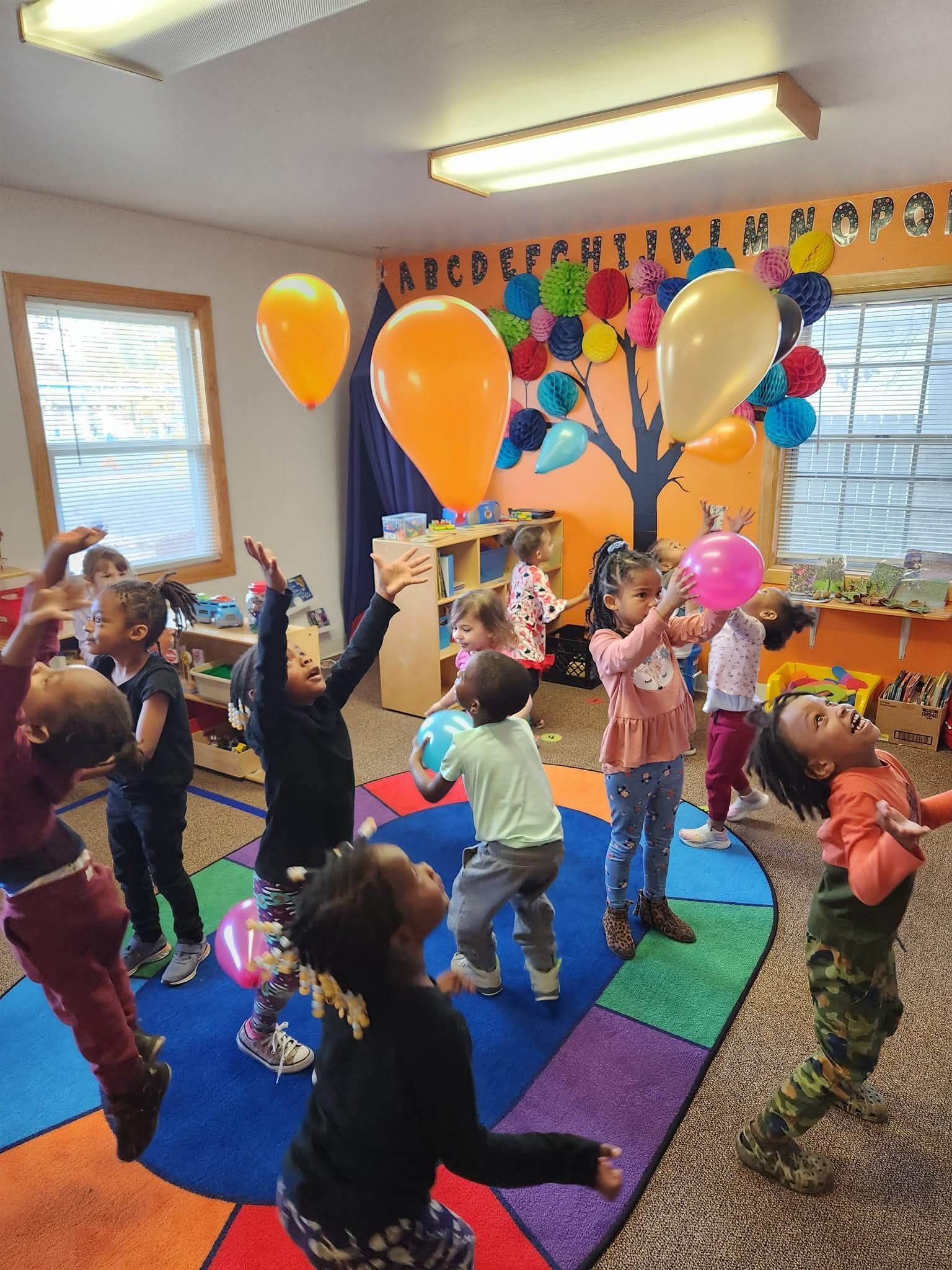 Children playing with colorful balloons in a brightly lit classroom with a vibrant, multi-colored rug.